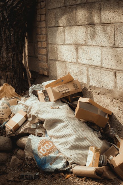 An outdoor scene depicting a pile of discarded packaging materials and waste, located at the base of a brick wall with visible mortar joints. The waste includes open cardboard boxes, some flattened and others partially expanded, along with large white sacks made of woven plastic material, which appear slightly crumpled and contain additional refuse. The packaging materials are in various shades of brown and tan, with some boxes displaying printed labels. The background features a rough brick wall with a visibility of mortar lines, and the ground consists of dirt and small rocks, suggesting an informal or neglected area. A dark tree trunk is partially visible on the left side of the image, indicating an outdoor environment. The lighting is natural, casting soft shadows and highlighting the textures of the cardboard and sacks, relevant to private waste handling or alternative rubbish removal services like those offered by [COMPANY_NAME]. This scene illustrates the type of waste accumulation that may be managed through independent rubbish collection or on-site clearance solutions, emphasizing the importance of proper waste disposal in non-urban settings.