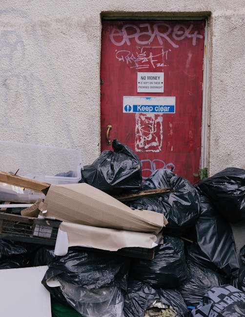 A collection of black garbage bags and discarded packaging materials stacked against a beige, textured wall beneath a red metal door covered in graffiti and stickers. The bags appear full and sealed, with some cardboard and plastic debris resting among them. The door, situated within a concrete or cement surface, features signs including a white notification saying 'NO MONEY, NOT OPEN OR CLOSED, PRIVATE OR ADVISED' and a blue sign with 'Keep clear.' The environment suggests an area for waste disposal outside a building, and the scene relates to independent rubbish collection or on-site clearance services, highlighting the importance of proper waste management handled by companies like rubbishremovalcrystalpalace.co.uk.