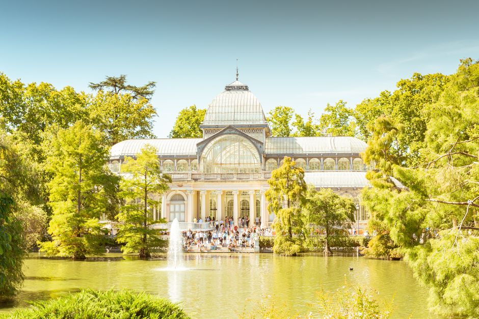 A large, ornate glass greenhouse with a domed roof and intricate architectural details, situated amidst lush green trees and foliage. In front of the greenhouse, there is a reflecting pond with a central fountain creating a vertical water spray. The pond's surface is calm, bordered by dense trees and shrubs with vibrant green leaves, some of which extend over the water. The greenhouse's glass panels are framed by light-colored stone or metal, and a crowd of people can be seen gathered on the steps leading up to the entrance, suggesting a popular visitor attraction. The bright, clear sky above indicates a sunny day, enhancing the natural light that illuminates the structure and surrounding landscape. This scene exemplifies an environmentally integrated setting adjacent to a site where waste removal or project clearance might occur, with the greenery providing a serene backdrop to the architectural feature.