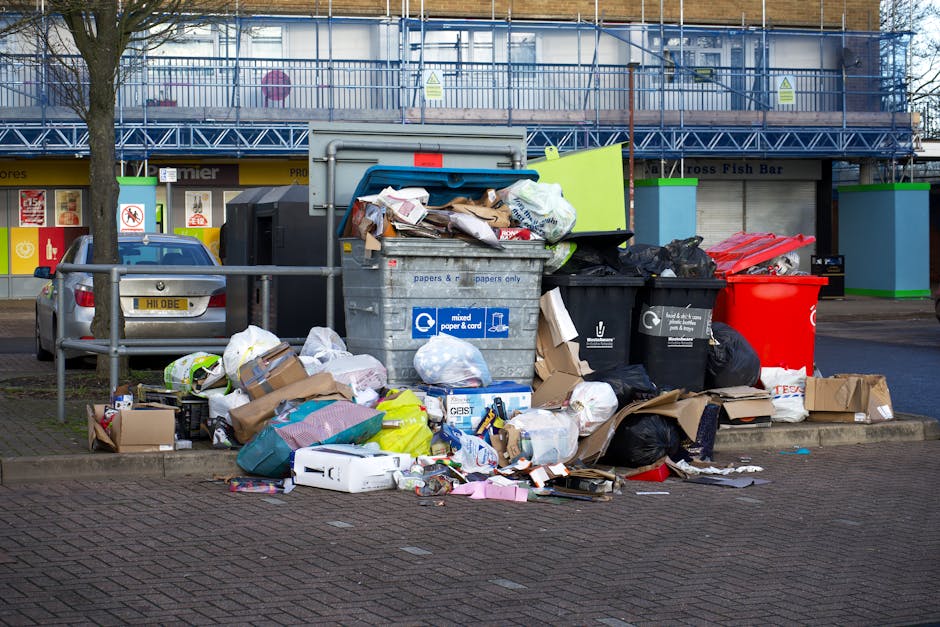 A collection of overflowing waste and rubbish bags located on a paved urban street scene, with various discarded items such as cardboard boxes, plastic bags, paper waste, and loose packaging piled around a large grey mixed waste bin, which is partially open and filled with crumpled paper, cardboard, and other refuse. The waste is spilling onto the surrounding pavement, some materials leaning against the bin or scattered on the ground. In the background, there are parked cars, including a grey vehicle directly behind the waste, as well as commercial shopfronts visible along the sidewalk, with signs and colorful window displays. Further behind, a multi-story building under construction is visible, covered with scaffolding and protective sheeting. Natural daylight illuminates the scene, highlighting the contrast between the clean construction site and the disorderly rubbish pile; this image illustrates the type of private waste accumulation that rubbish removal services like those offered by [COMPANY_NAME] can manage, emphasizing the importance of timely rubbish disposal and on-site clearance for urban environments.