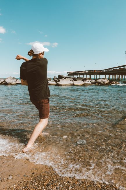A woman with curly hair tied back, wearing a white cap, a dark short-sleeved shirt, and brown shorts, standing in shallow water at a pebble beach. She is facing away from the camera, with her right arm extended across her chest and her left arm bent, creating a relaxed stance. The water is clear and gently lapping at her ankles, revealing smooth, rounded pebbles and small stones beneath. In the background, a line of large rocks separates the shoreline from deeper water, with a concrete pier or bridge extending outward supported by pillars, and a partly cloudy blue sky above. The scene conveys a calm, coastal environment, typical of outdoor leisure or shoreline activities. The environment appears bright and sunny, with natural lighting highlighting the textures and colors of the rocks, water, and sky, offering a natural connection to outdoor related activities which could relate to the context of waste management or environmental considerations, though the scene itself shows no rubbish or waste materials.