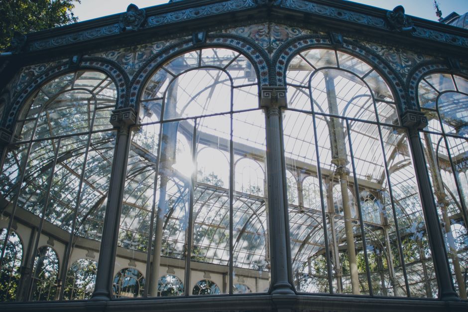 A detailed view of an ornate, vintage-style glass and metal greenhouse structure with large, arched windows and decorative columns. The glass panels are framed with slender metal bars, allowing sunlight to filter through and casting bright reflections on the surfaces. The intricate metalwork features decorative patterns and moldings along the top edges of the arches, with some visible weathering in the finish. Inside and behind the glass, a glimpse of lush greenery and trees can be seen, suggesting the greenhouse is situated in a garden or park setting. The sunlight creates a bright, airy atmosphere highlighting the texture of the metal and the transparency of the glass. The structure appears well-maintained with a classic design, possibly serving as a botanical conservatory or decorative greenhouse, with no signs of rubbish or waste, reflecting a pristine environment suitable for conservation or private horticultural use. This scene emphasizes the aesthetic and structural details of the greenhouse, aligning with services that focus on on-site clearance and preservation of such intricate structures.