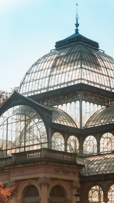 A detailed close-up of a historic glass and iron dome structure showcasing multiple curved glass panels with metal framing, sitting atop a classical-style building. The domes vary in size, with the largest centrally positioned and smaller ones surrounding it, all featuring intricate metal detailing and decorative finials at their peaks. The glass panels are transparent, revealing the internal iron supports, and have a slightly reflective surface that captures the soft daylight, contributing to a bright and airy appearance. The structure is set against a light blue sky, with the warm tones of the building's stone base visible at the bottom. The surroundings suggest an outdoor setting, possibly part of a conservatory, greenhouse, or historic public building, reflecting traditional Victorian or Edwardian glasswork designs. This scene visually aligns with the context of private or independent collection and on-site clearance for historic or ornamental glass structures, fitting within the scope of rubbish removal or alternative waste handling services by companies like rubbishremovalcrystalpalace.co.uk.