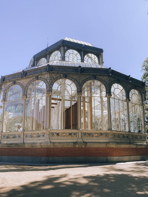 A large glass greenhouse structure with a curved roof exhibits a detailed framework of metal and glass panels, reflecting a mix of transparency and textured finishes. The building is situated outdoors amidst bare trees, with the silhouette of branches visible against the early evening sky, which features soft, diffused light and some cloudy patches. The greenhouse's transparent glass panels reveal an interior that appears to contain plants or gardening elements, although details are obscured by the external grid-like framework. The structure's metallic framing has a dark finish, contrasting with the lighter, reflective surfaces of the glass, which pick up ambient natural light. Surrounding the greenhouse are natural elements, including the silhouettes of trees with sparse foliage, indicative of late autumn or early winter. The scene captures a quiet, atmospheric moment, emphasizing the building's architectural complexity and the natural environment, aligning subtly with themes of private or alternative waste handling by showcasing an unconventional and potentially expansive structure that may be associated with a botanical or horticultural setting, often requiring specialized waste or material management as part of site maintenance or renovation projects.